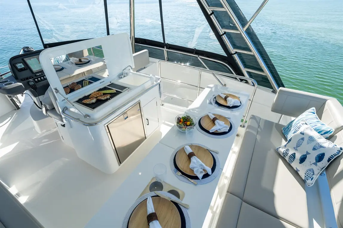 A luxury yacht features a grill with cooked food, a dining table set for four, and a scenic ocean backdrop.